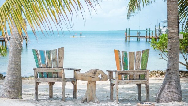 Zwei rustikale Holzstühle mit bunten Kissen an einem sandigen Strand, im Hintergrund eine Palme und eine Holzstege.