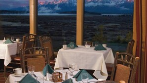 A dining area with tables set for two, large windows offering a view of snow-capped mountains and a lake. - Charming Jackson Hole cabin bordering Grand Teton National Park (Jackson)
