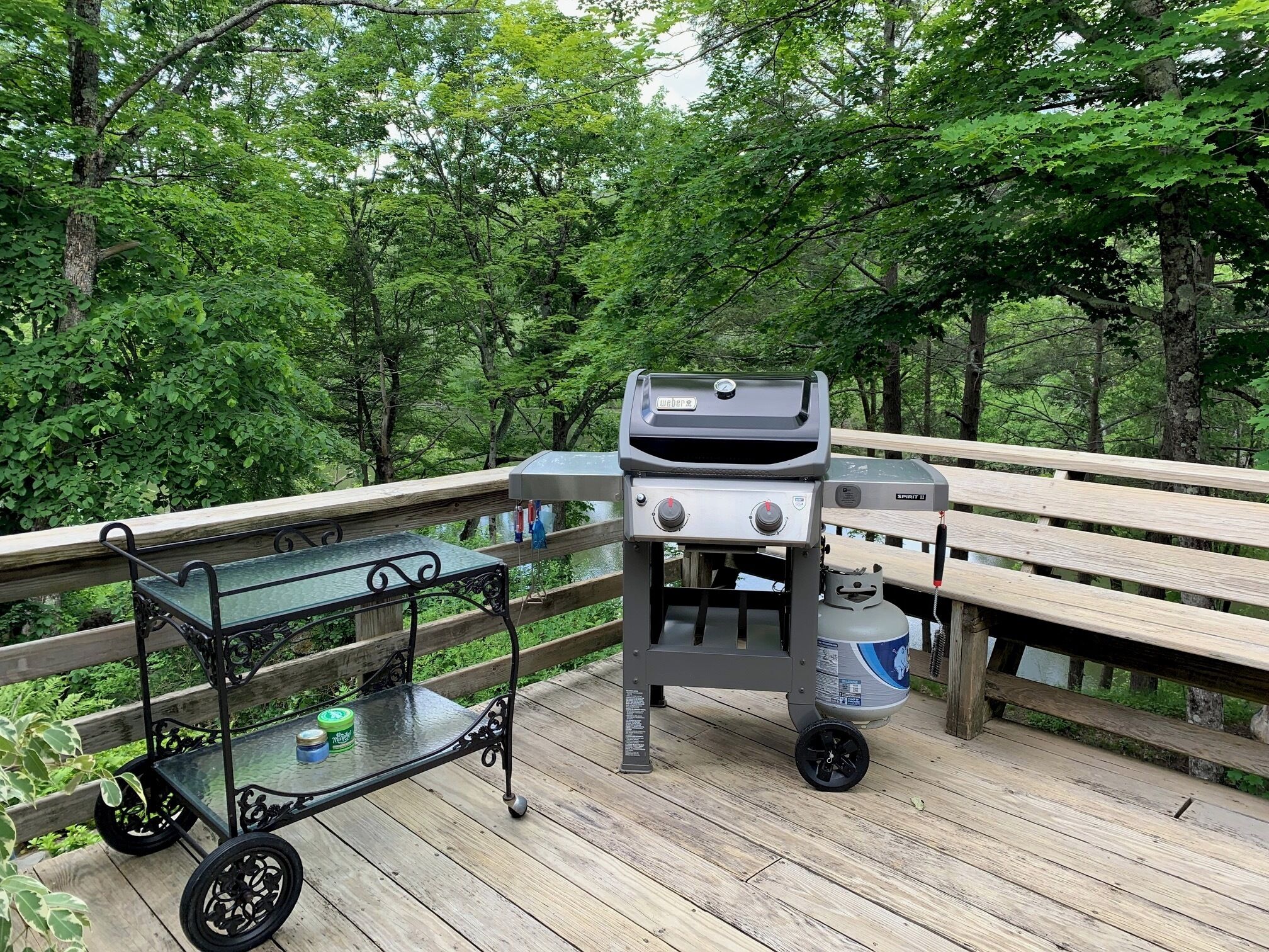 A portable Weber Spirit II grill on a wooden deck with a glass table and a cart.