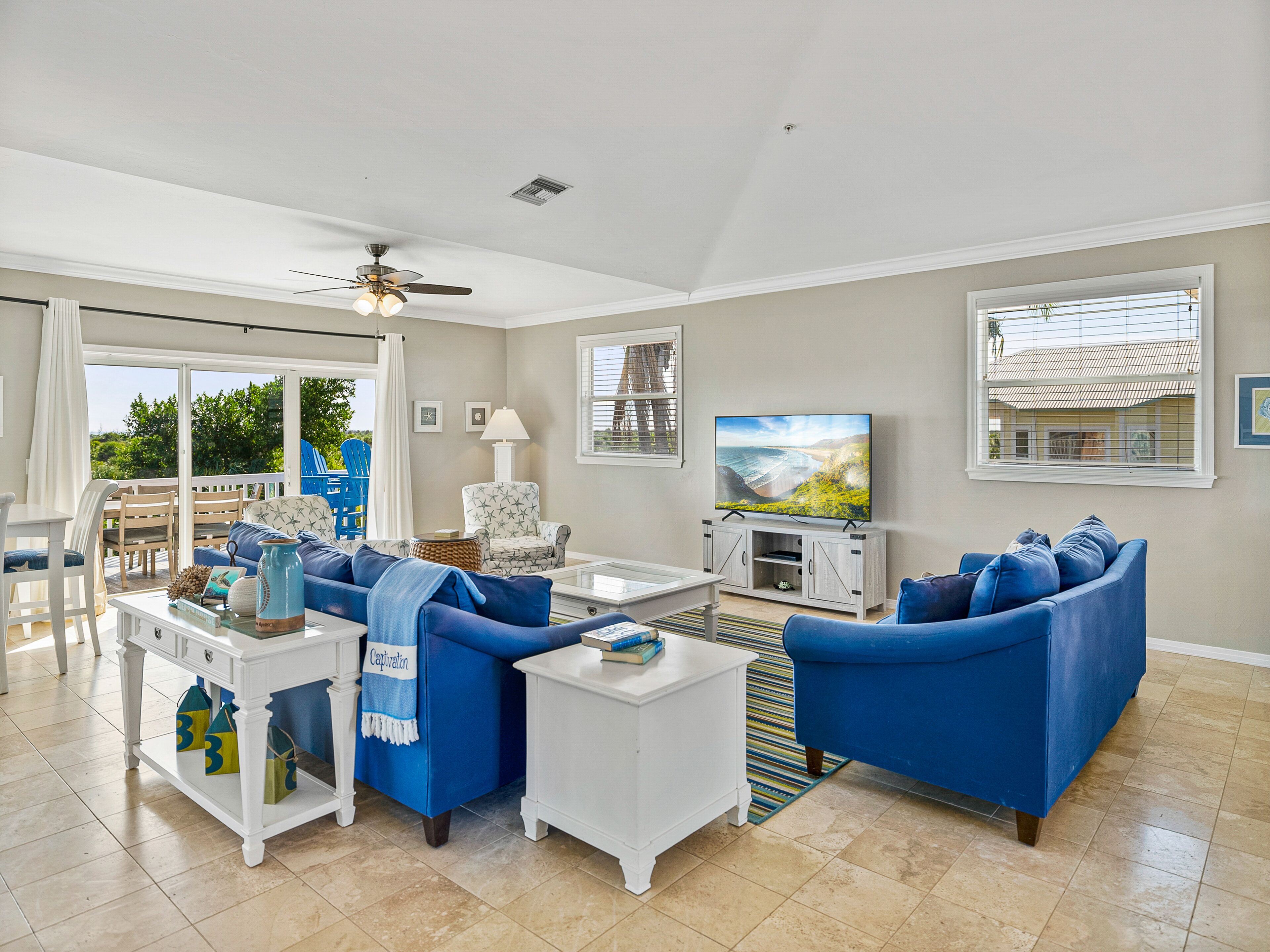 A spacious living room with a large TV, blue sofas, a white coffee table, and a ceiling fan.