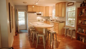 A kitchen with wooden cabinets, a central island with stools, a refrigerator, and a window with a view of trees. - Cedar Point Cottage- Perfect Lake Michigan Retreat (Mears)