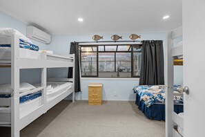 A dormitory room with bunk beds, a desk, a window with curtains, and an air conditioning unit. - ACCOMMODATE 19 -  OCEAN FRONT. (Pukehina)