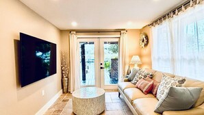 Un salón con sofá, televisor de pantalla plana, una mesa redonda y vistas al exterior a través de un ventanal.