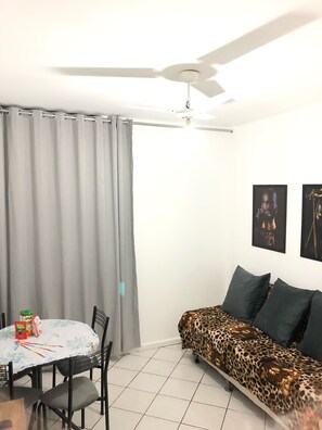 Un salon avec un canapé, une table à manger et des chaises, un ventilateur de plafond et une fenêtre avec des rideaux.