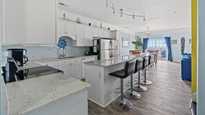 Une cuisine moderne avec des meubles blancs, un îlot central doté de tabourets de bar, un réfrigérateur en acier inoxydable et un coin repas avec un canapé bleu et des chaises en bois.