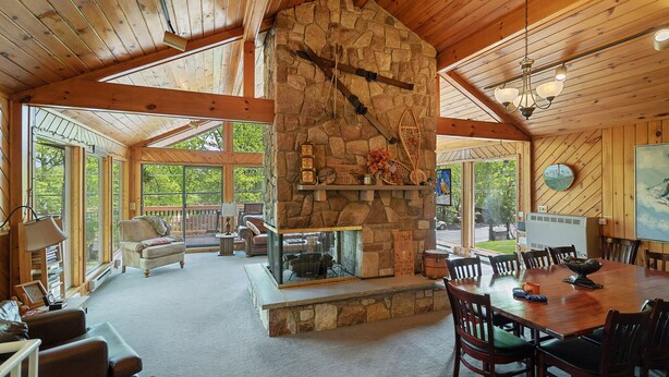 A spacious room with a stone fireplace, wooden ceiling beams, a dining table with chairs, and large windows offering a view of the outdoors. - Repeat Retreat — Your Place to Come Back To! (Lake Harmony)