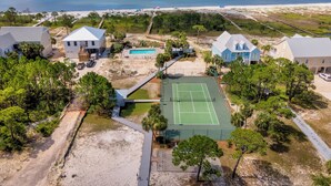 Vue aérienne d’un quartier résidentiel situé sur la côte, avec des maisons, un court de tennis et une plage.