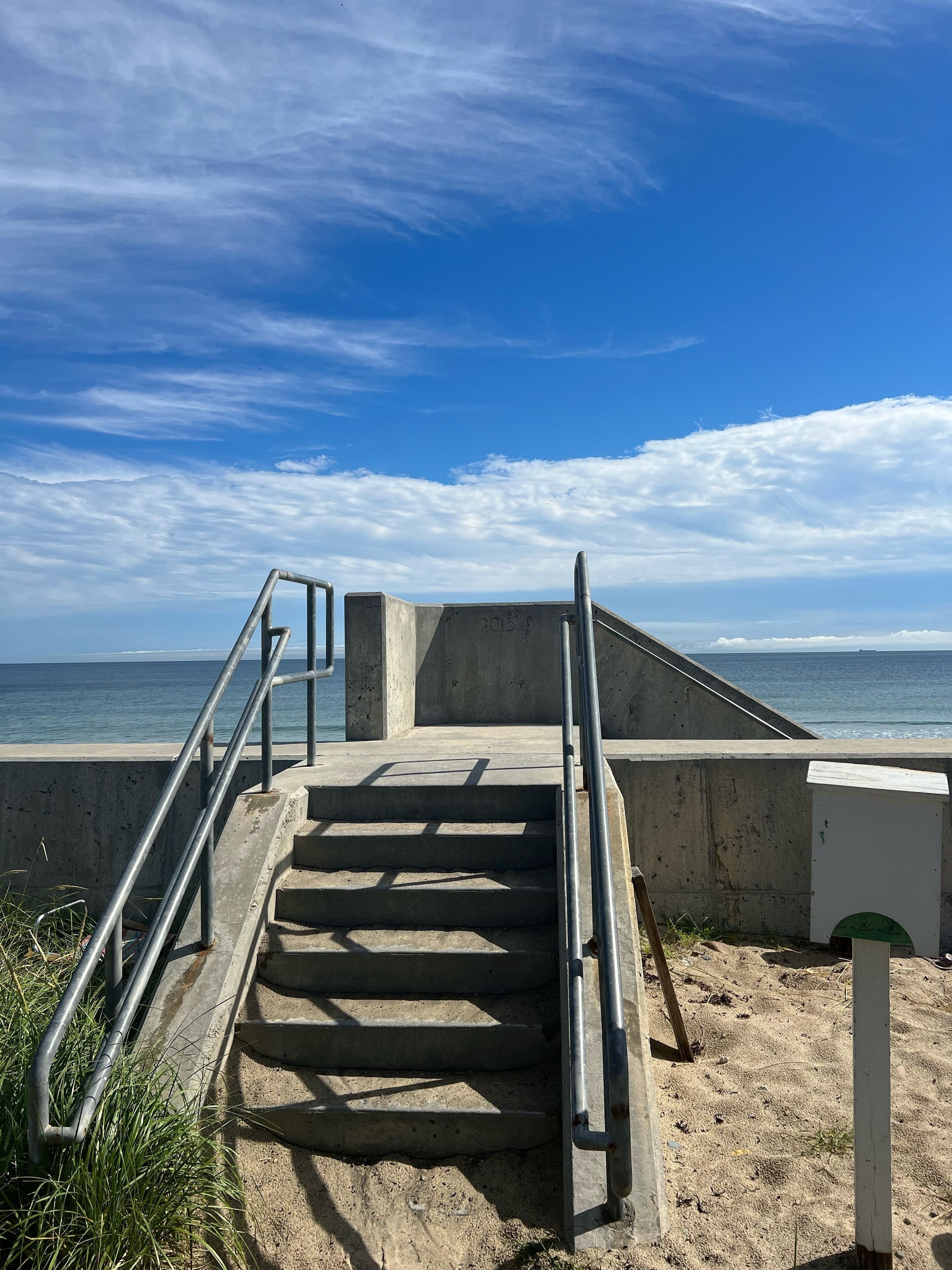 Deguts de formigó que baixen a una platja, amb baranes de metall, un mur de formigó i una vista clara de l'oceà.