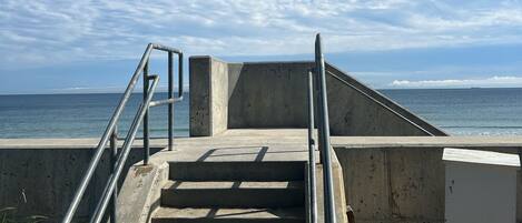 Deguts de formigó que baixen a una platja, amb baranes de metall, un mur de formigó i una vista clara de l'oceà.