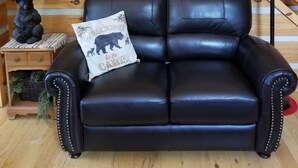 A black leather sofa with a bear-themed pillow, a lamp, a framed picture, and a wooden wall with a circular mirror. - Papa Bear's River Cabin- 3 bedroom, 2 full bath, Riverfront (Shenandoah)