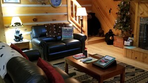 A cozy living room with a leather sofa, a coffee table, and a fireplace. There is a clock on the wall and a star decoration above it. - Papa Bear's River Cabin- 3 bedroom, 2 full bath, Riverfront (Shenandoah)