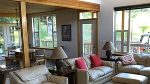 A spacious living room with high ceilings and wooden beams, featuring a beige leather sofa set, a coffee table, and a dining area with a table and chairs.