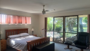 Une chambre avec un lit, une table de chevet, une lampe, un ventilateur de plafond et une vue sur l’extérieur, accessible par une porte-fenêtre coulissante.