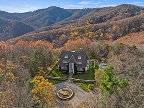 A large house with a well-maintained lawn and a circular driveway, surrounded by autumn-colored trees and hills in the background. - Ultimate Boone/Blowing Rock Luxury Retreat: Pool, Hot Tub, Fire Pit & Epic Views (Boone)