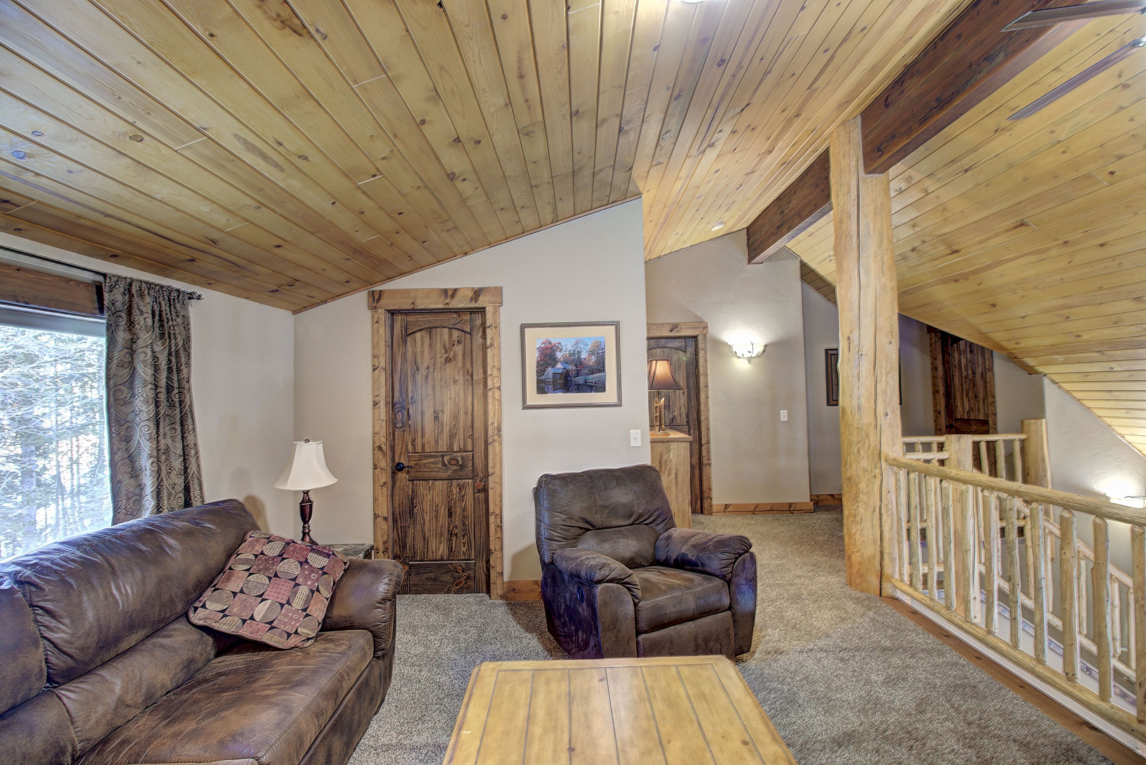 Una sala de estar acogedora con un sofá de cuero marrón, un sillón a juego y una mesa de centro de madera. Se ve una puerta de madera y una escalera al fondo.