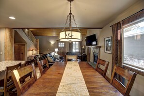 Uma sala de jantar com uma longa mesa de madeira, cadeiras, um candelabro e uma televisão na parede.