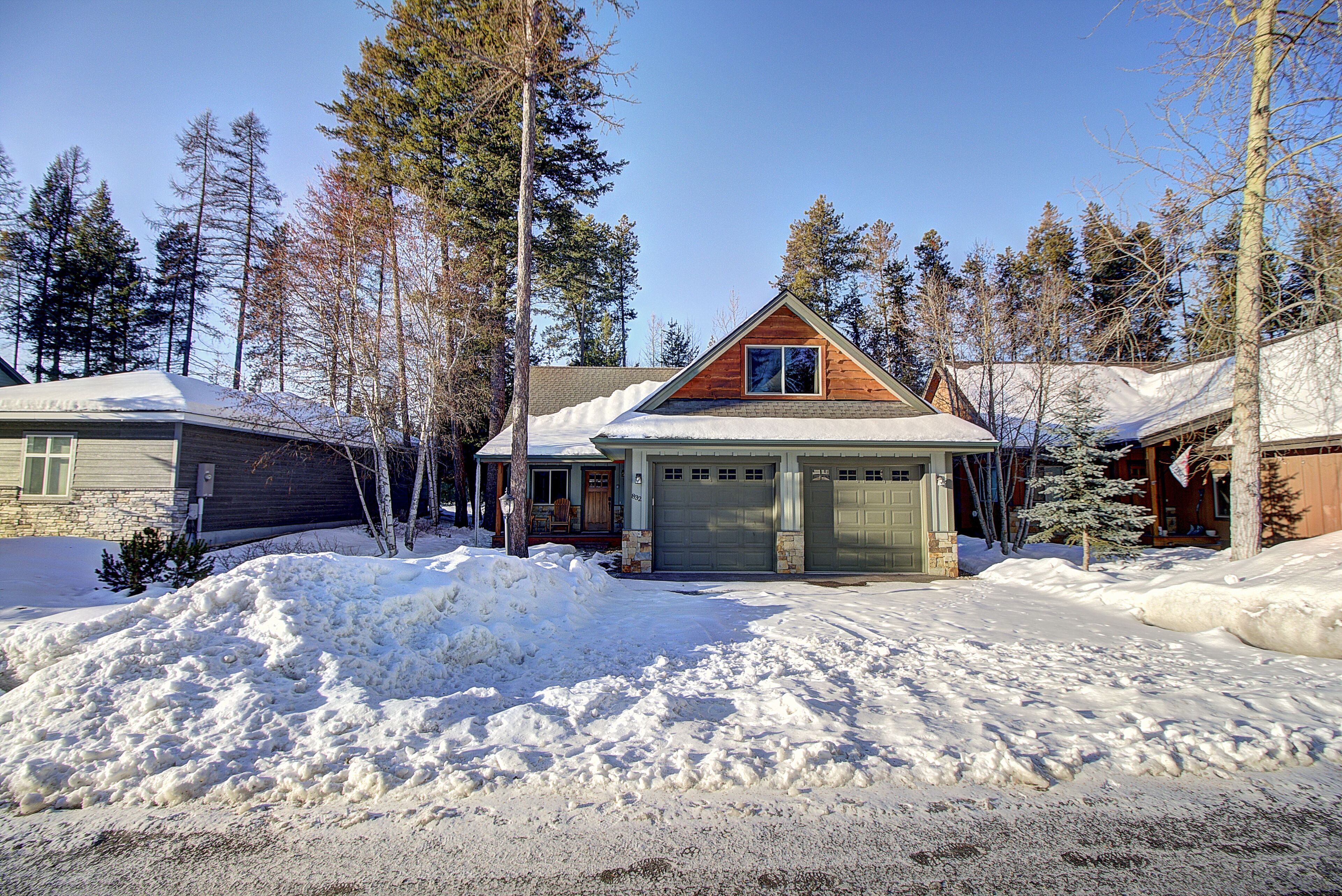 Une cabane en rondins à deux étages, dotée d’un garage, entourée d’un paysage enneigé et de grands pins.
