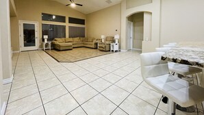 Ein geräumiges Wohnzimmer mit einem Ecksofa, einem Esstisch mit Stühlen, einem Deckenventilator und großen Fenstern mit Jalousien.