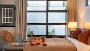 Una camera d'hotel con una grande finestra, un letto con coperta trapuntata, una sedia, un tavolo e un quadro appeso alla parete.