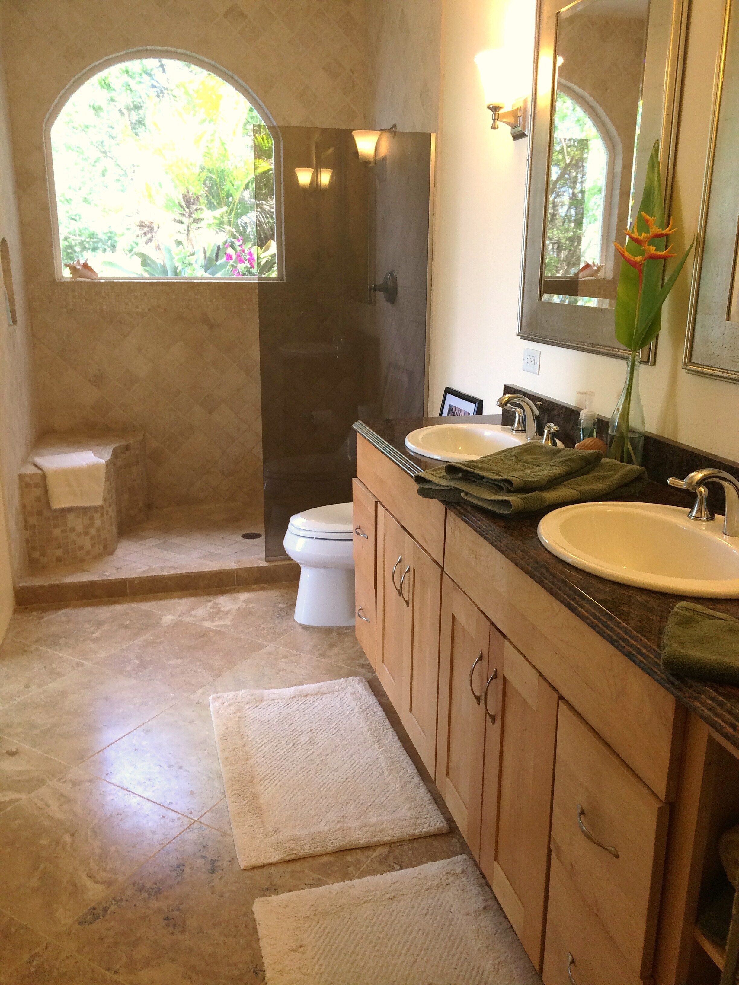 A bathroom with two sinks, a toilet, and a shower. There is a window with a view of greenery outside.