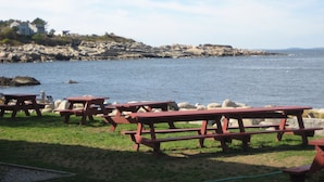 A coastal picnic area with wooden tables and benches, a rocky shoreline, and a view of the sea.