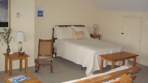 A bedroom with a bed, a chair, a nightstand, a lamp, a table, and a framed picture on the wall.