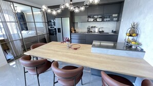 Moderni keittiö, jossa on keskeinen saareke, ruokailutila puupöydällä ja nahkaisilla tuoleilla sekä näkyvillä oleva keittiötaso, jossa on erilaisia kodinkoneita ja hedelmäkori.