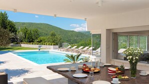 Zona de comedor junto a la piscina con vista a la montaña, con mesas al aire libre y un sistema de buffet.