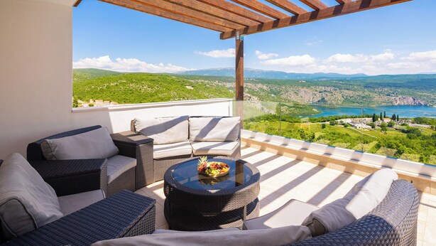 Un balcón con mobiliario de mimbre, una mesa redonda con frutero y vistas a colinas verdes y un lago.