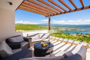 En balkon med fletmøbler, et rundt bord med en skål frugt, og udsigt til grønne bakker og en sø.