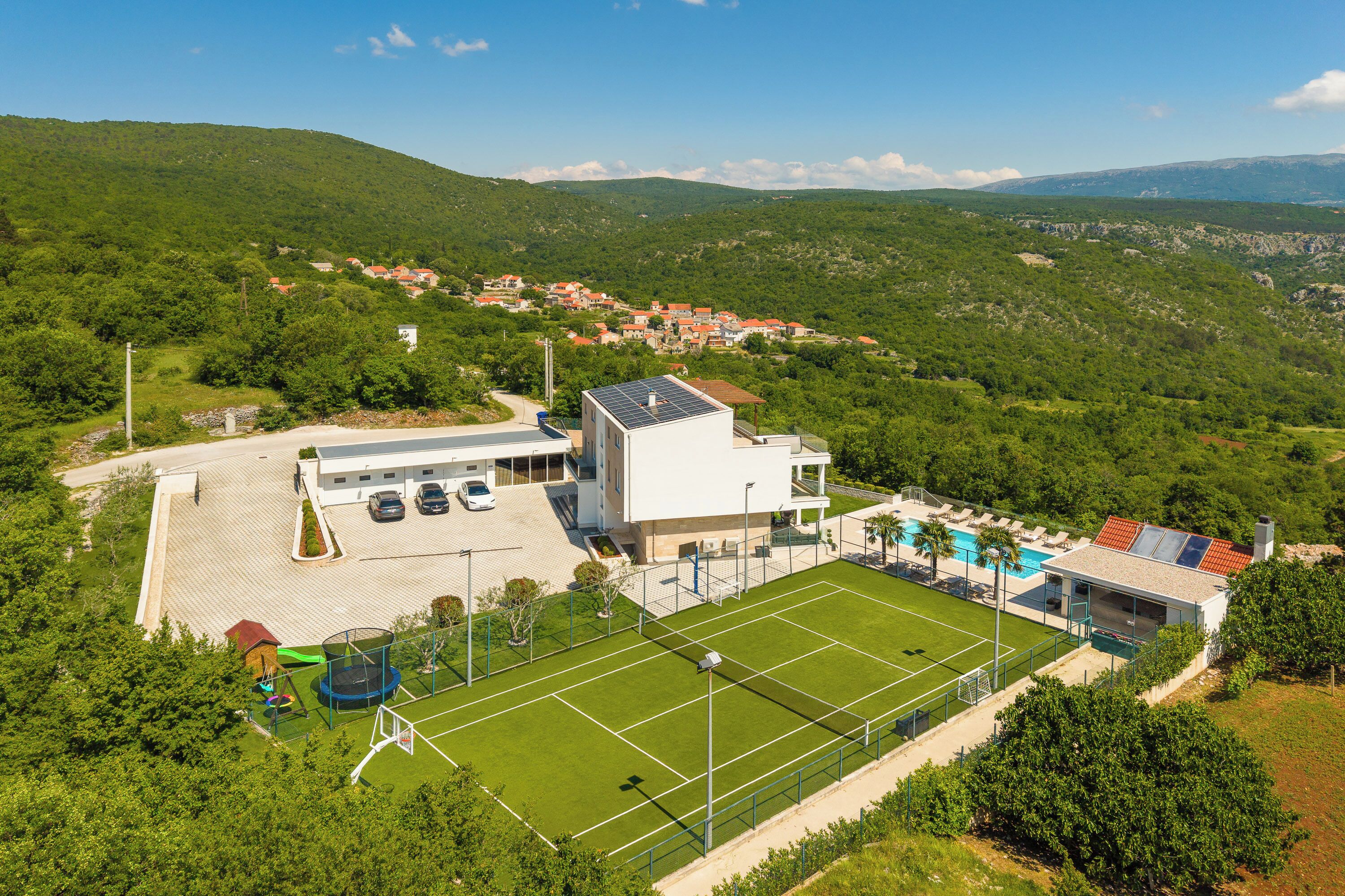 A modern building with a tennis court, surrounded by lush greenery and a hilly landscape.