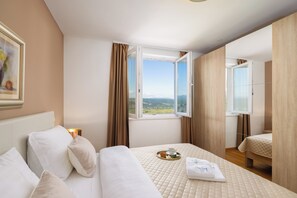A hotel room with a bed, a window with a view, a bedside table, and a painting on the wall.
