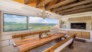 Una mesa de comedor de madera con bancos, una araña, una chimenea y ventanas amplias con vistas al paisaje.