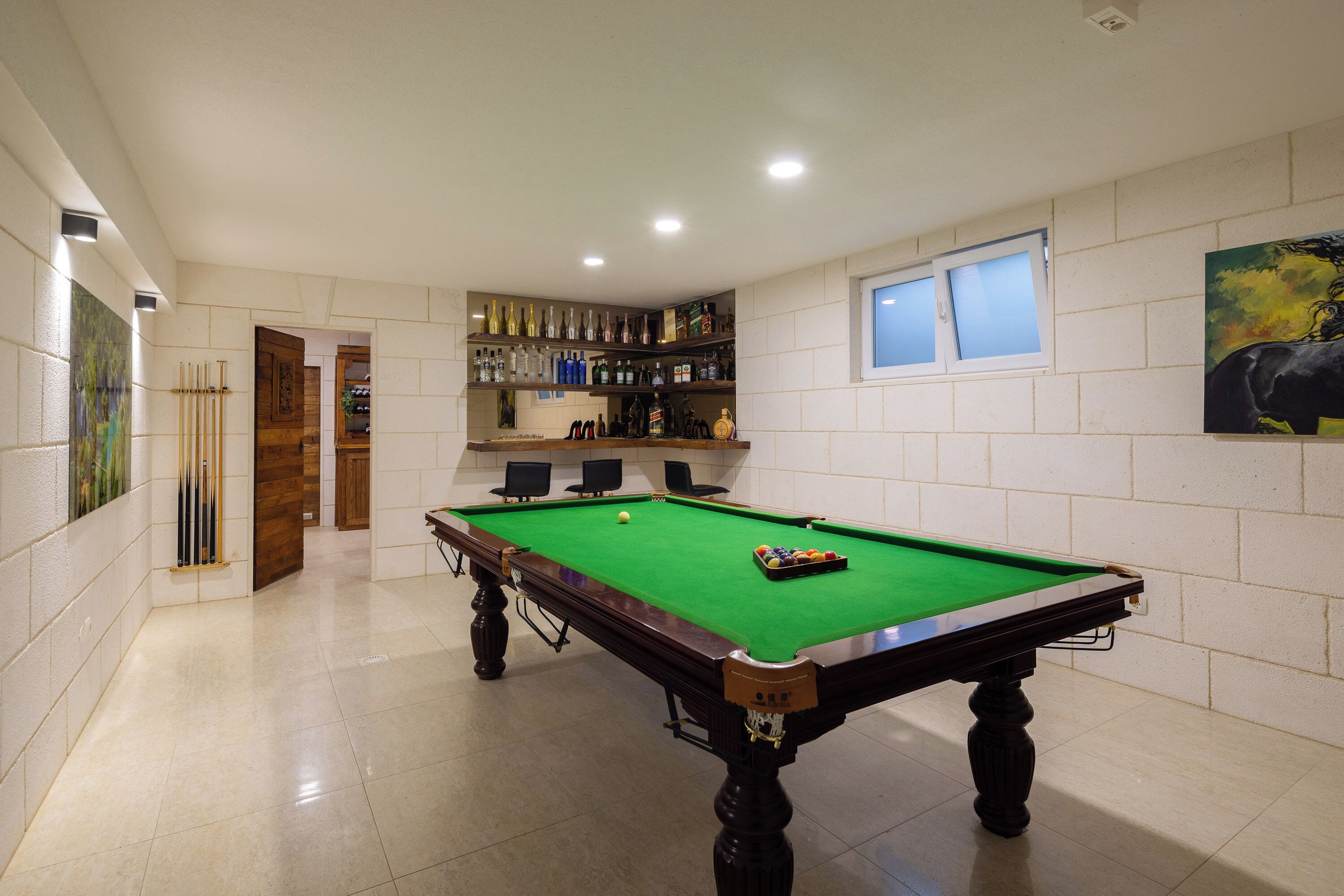 Une table de billard recouverte d’un drap vert, entour ée d’un sol carrelé de carreaux blancs, et une étagère contenant diverses bouteilles et verres.
