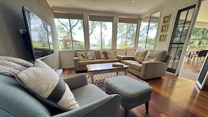 Amplia sala de estar con sofá, reposapiés y mesa de centro, ventanas grandes con vistas a árboles y una puerta corrediza de vidrio que da a un espacio exterior.