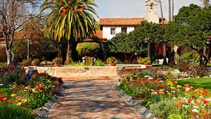 Un camí al jardí que condueix a un edifici amb una torre de campanars, envoltat de diverses plantes i flors.