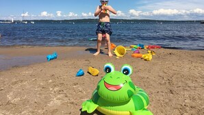 Escena de playa con un niño jugando con juguetes y un flotador con forma de rana sobre la arena.