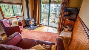 Ein gemütliches Wohnzimmer mit Kamin, Ledersofas, Fernseher und einem großen Fenster mit Blick auf einen Wald.