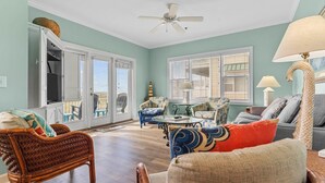 A spacious living room with wicker furniture, a ceiling fan, and large windows with blinds. - CottageVacations4u "Ocean Paradise" -Ocean views! -pool club-dog friendly- elevator (Ocean Isle Beach)