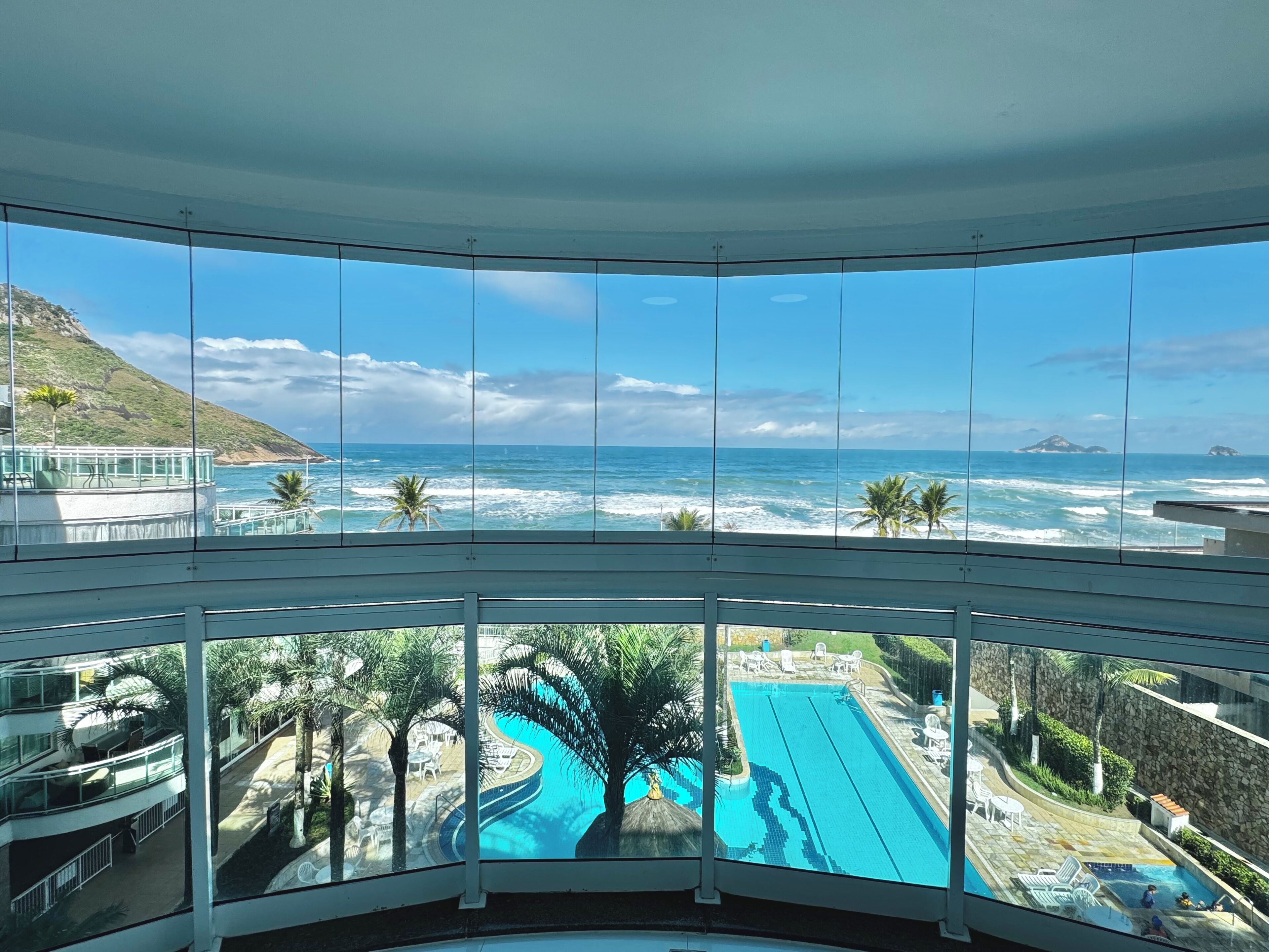 A view from a balcony overlooking a swimming pool and ocean, with palm trees and a mountainous landscape in the background.
