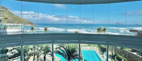 A view from a balcony overlooking a swimming pool and ocean, with palm trees and a mountainous landscape in the background.