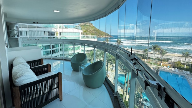A balcony with a view of the ocean, a pool, and a modern building in the background. - Pontal Beach Ocean View (Rio de Janeiro)