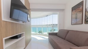 A modern living room with a large window overlooking a beach, a flat-screen TV mounted on a wooden panel, and a beige sofa. - Pontal Beach Ocean View (Rio de Janeiro)