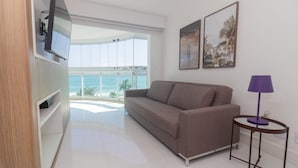 A modern living room with a sofa, a TV mounted on a wooden panel, a small table with a lamp, and two framed pictures on the wall. - Pontal Beach Ocean View (Rio de Janeiro)