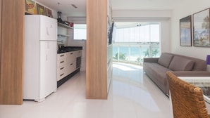 A modern kitchen with white appliances, a dining area with a wicker chair, and a balcony with a view of the ocean. - Pontal Beach Ocean View (Rio de Janeiro)