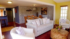 Un salon avec un canapé à rayures, un fauteuil blanc et une table basse en osier. On y trouve également un coin repas avec des chaises en bois et une table, ainsi qu’une cuisine équipée d’un réfrigérateur et d’un tabouret en bois.