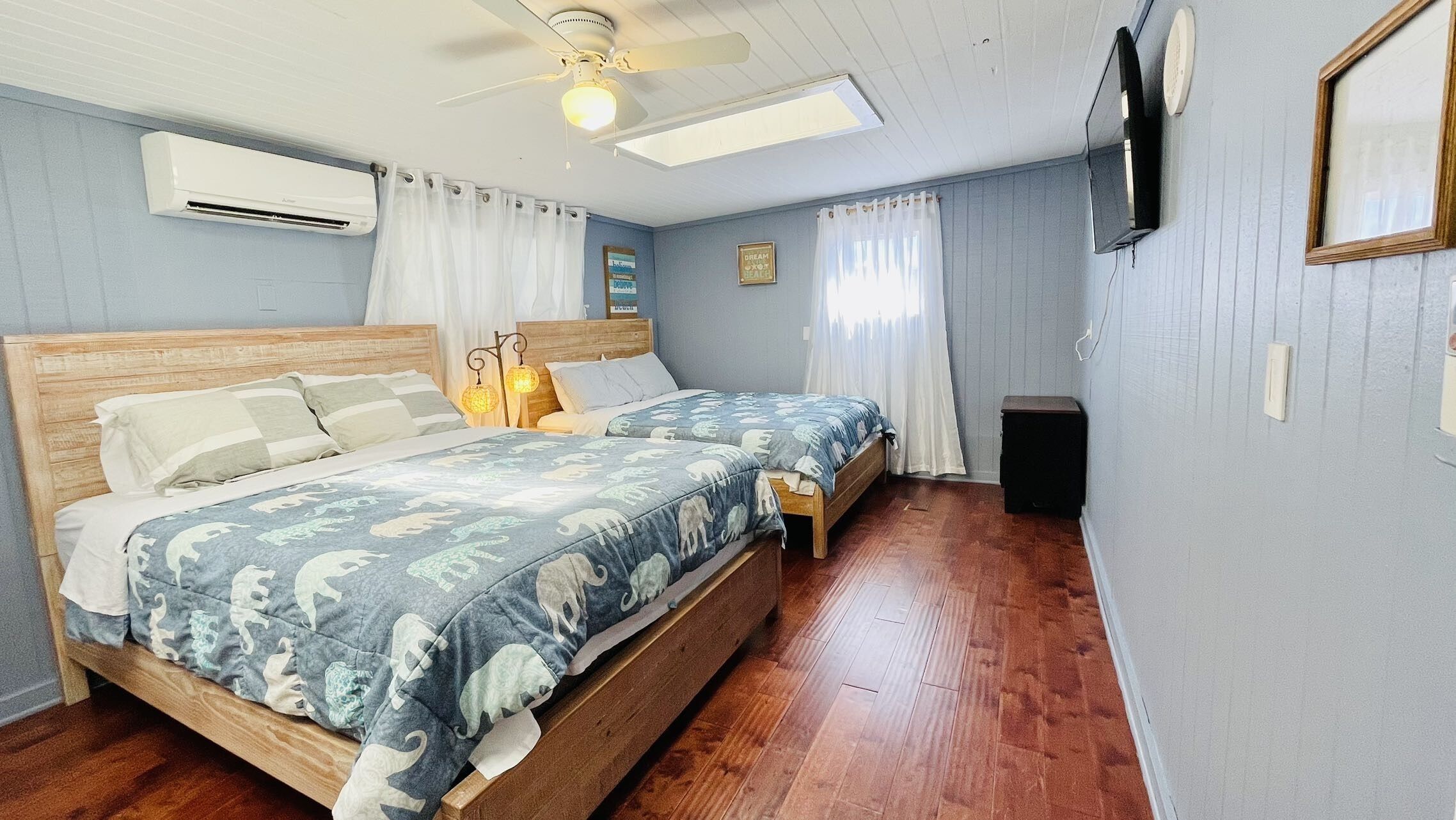 Une chambre à coucher avec un lit en bois, deux tables de chevet, un ventilateur de plafond, un appareil de climatisation, un téléviseur fixé au mur et une fenêtre avec des rideaux.