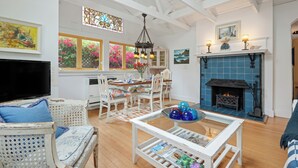 A spacious living room with a fireplace, white furniture, and a dining area with a view of pink flowers through the windows. - Seaside Cottage - Romantic Haven by the Beach (Montecito)