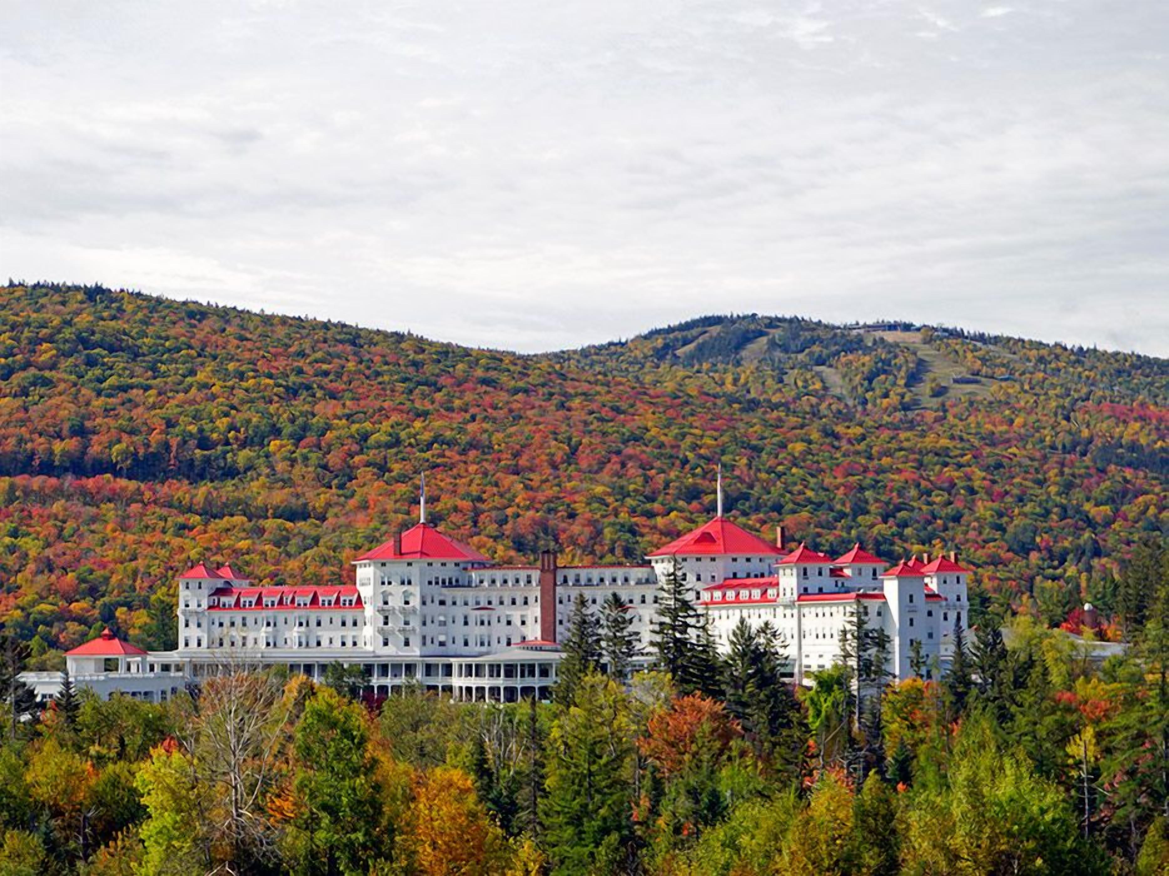 Un hotel grande de varios pisos con techos rojos, rodeado de árboles y colinas de colores otoñales.