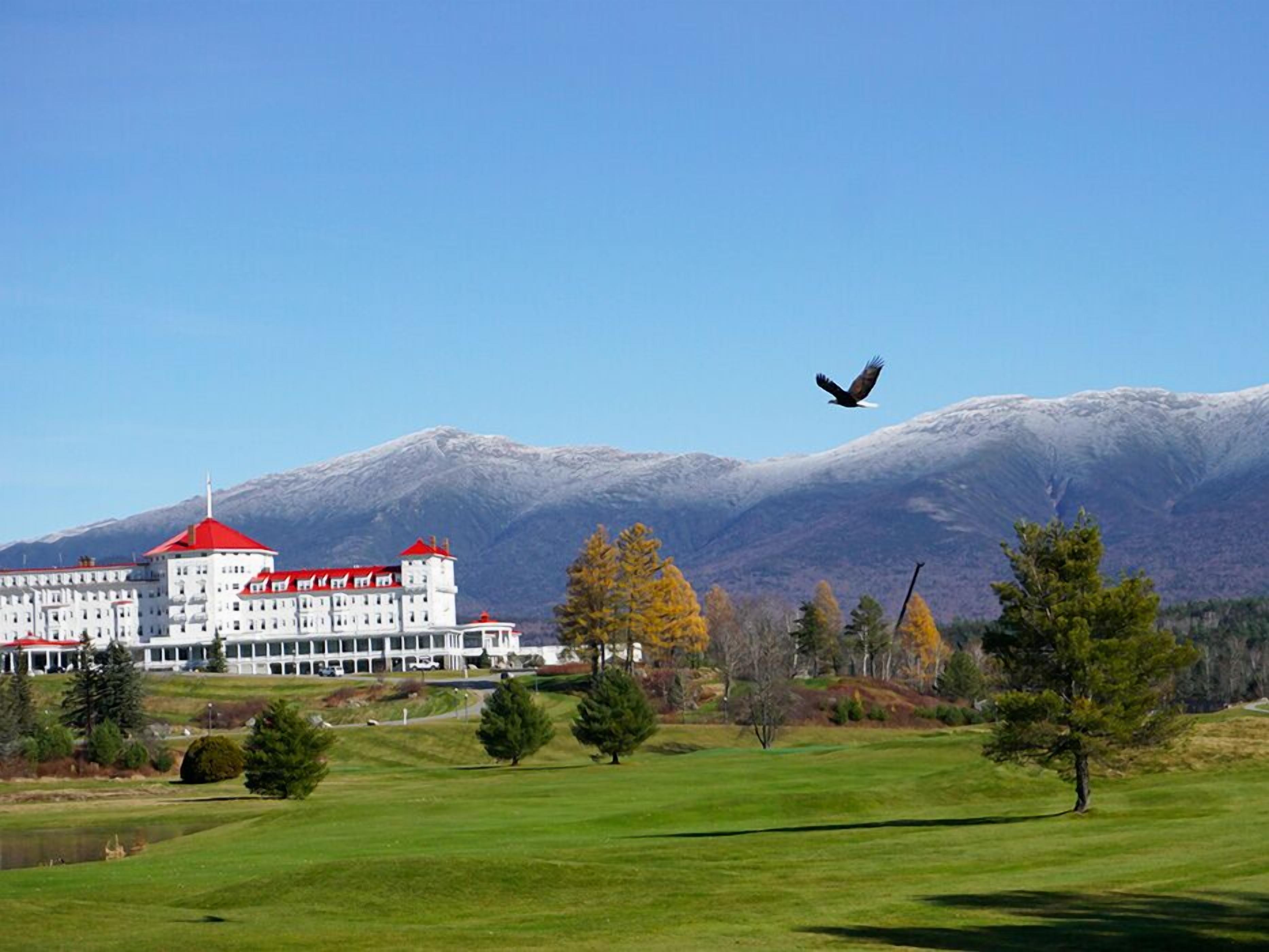 A large building with a red roof, surrounded by a green landscape and mountains in the background.